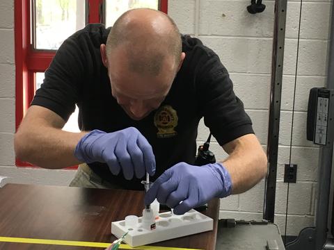 A man in a black shirt labeled "Fire Marshal" is wearing plastic gloves as he bends over a vial in a holder on a table. 
