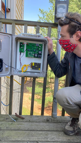 A prototype low-cost GHG sensor box opened with the circuits showing and Tyler Boyle crouched next to it on a deck at NIST