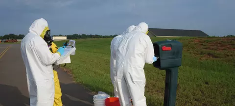 Four figures in coveralls stand outdoors around a mailbox, looking at a clipboard and other items. 