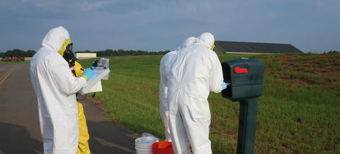 Four figures in coveralls stand outdoors around a mailbox, looking at a clipboard and other items. 