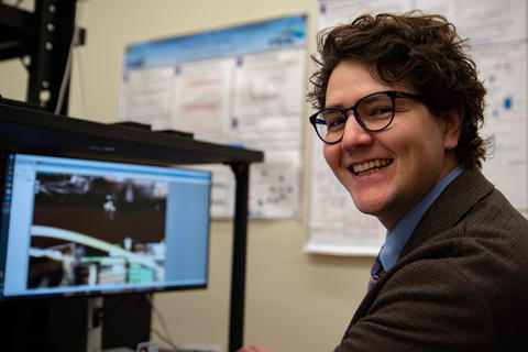 Liam Connolly sits next to a computer monitor and smiles for a photograph.