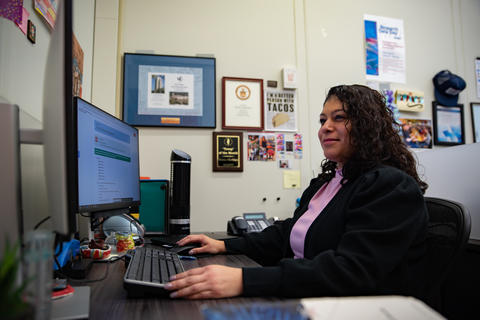 Serena Martinez sits at a computer workstation in an office setting. 