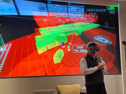 A man wears a virtual reality headset in front of a large projection screen with red and green graphics.