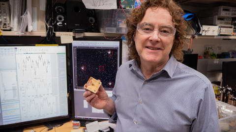 NIST's Andrew Wilson holds an ion trap in his lab in Boulder, Colorado. Behind him are screens depicting dots. 