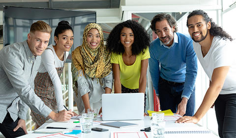 Workforce photo showing group of diverse employees standing at a table with a laptop.