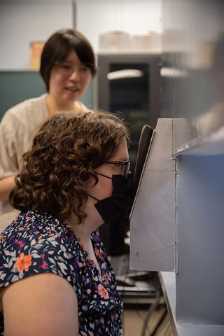 Megan King wears a black mask as she looks into a lighting test device, with Jane Li standing in the background. 