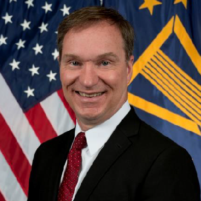 Headshot of a man in a suit standing in front of the American flag. 