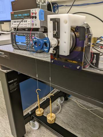 Scientific equipment on a lab table includes stacked boxes with readouts and device with two weights suspended by wires.