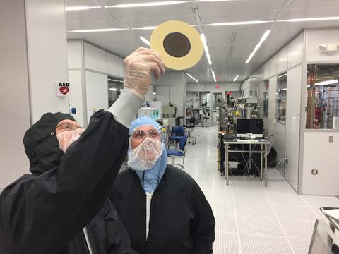 Two people wearing coveralls in a "clean room" lab look up at a disk-shaped object that one of them is holding. 