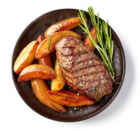 Overhead view of a dinner plate with a grilled steak, potatoes and a green garnish. 