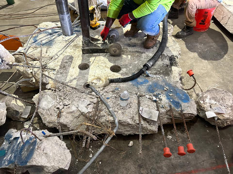 A slab of concrete with ragged edges sits on a warehouse floor as researchers use equipment to extract core samples. 