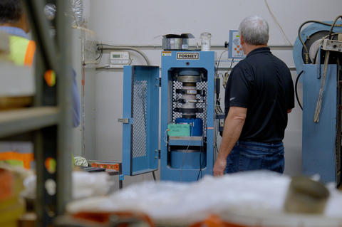 A researcher faces away from the camera toward a concrete core sample held vertically in a blue metal testing machine. 