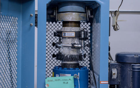 A concrete core sample shatters as it is held vertically inside a blue metal testing machine. 