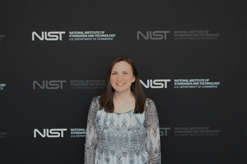 A woman with long brown hair wearing a white and gray blouse, standing in front of a NIST photo backdrop
