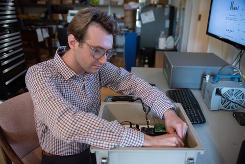 A man wearing safety glasses and sitting at a computer workstation reaches into a box with wiring and circuitry.