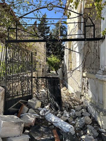 A narrow alley, open to blue sky above, is blocked with rubble and broken iron gates.