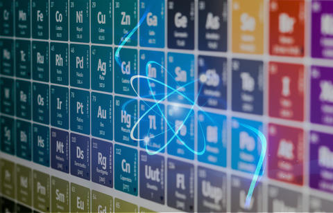 Illustration  with a stylized representation of an atom with electron orbits superimposed on a colorful periodic table of the elements. 