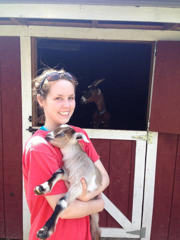 A young Jerilyn Izac stands in front of a red barn holding a baby goat while a larger goat looks on from inside the barn. 