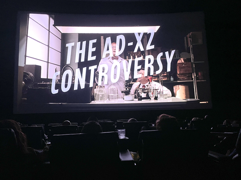 A darkened movie theater with a crowd looking at the screen. The screen shows the opening sequence of "The AD-X2 Controversy" with the words "The AD-X2 Controversy" on the screen.