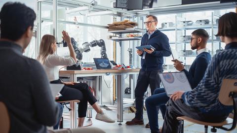 Several college-age people sit in a modern high-tech classroom or office with a table holding a robot arm and an older man standing with a tablet computer. 