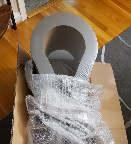 A partially opened cardboard box on the floor of a home contains a curved cat scratching post in shrinkwrap and bubble wrap. 