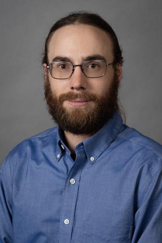 Portrait of Orion Kafka - an individual with brown hair, a mustache, beard, and glasses, wearing a blue collared button up shirt.