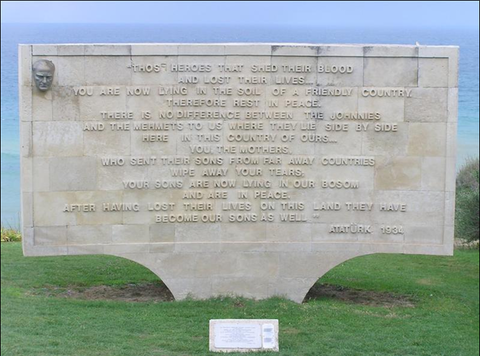 A white stone memorial displaying a long quote from Ataturk stands near the edge of a cliff by the water. 