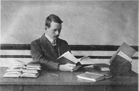 Historical photo shows Henry Moseley sitting at a desk reading a thick book, with other open books on the desk. 