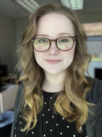 Woman standing in office with long wavy blonde hair, glassses, and a closed mouth smile. She is wearing a a gray cardigan over a black with white dots shirt.