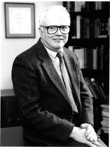 Vertical, black and white image of a man in a suit sitting at the edge of a desk.