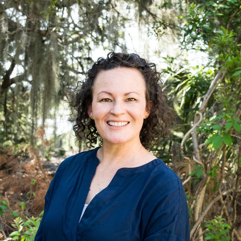Headshot of Carolyn Q Burdette. She has shoulder length, dark curly hair, fair skin, a big smile, and squinty eyes. She is wearing a dark blue blouse and is standing in front of spanish moss covered trees with Charleston Harbor marshlands behind that. 