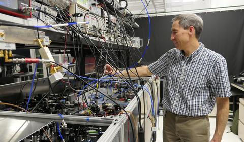 Tasshi Dennis stands in his lab and reaches toward a countertop full of equipment with lots of wires and circuitry.