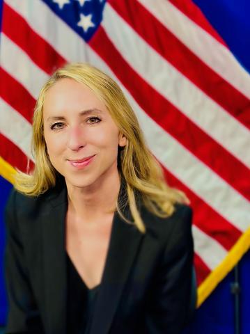 Woman with blond hair, black shirt and black jacket with a U.S. flag background