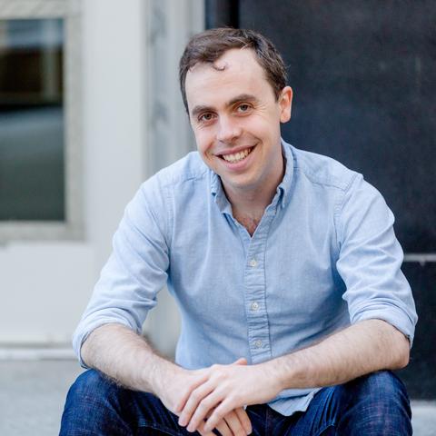 Man with dark hair sitting down, elbows on knees, wearing a blue shirt with sleeves rolled up 