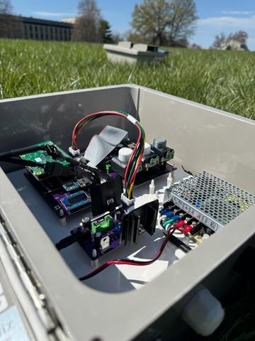 An open gray box filled with circuitry, wires, sensors and a single-board computer sits outside on a lawn.