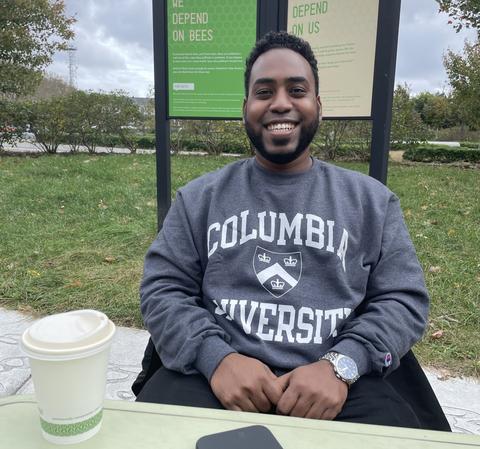 A man with short black hair, wearing a grey Columbia University sweatshirt and a watch, sitting at a table outside.