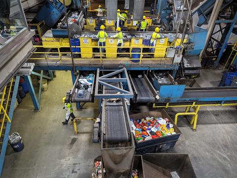 Workers at a recycling facility sort and separate recycled plastics into individually-labeled yellow bins. 