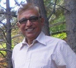 Headshot of a man with short light brown hair and glasses, wearing a button up collared shirt, standing outside in front of trees.