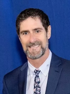 Headshot of a man with dark hair, beard and mustache wearing a suit and tie.