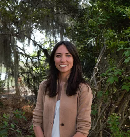 A woman with shoulder length black hair wearing a tan blazer and white shirt standing in front of a marshland.