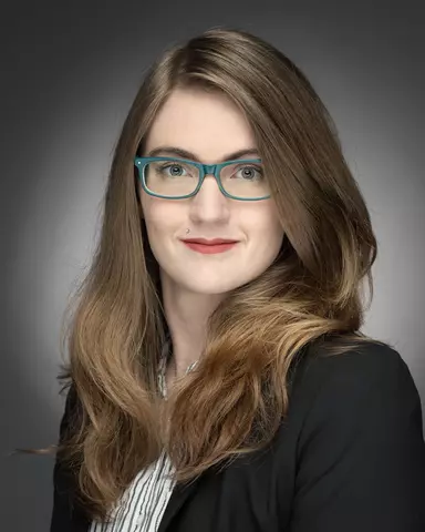 Indoor studio headshot of a woman with long, dark blonde hair in professional attire and glasses on a soft grey background.