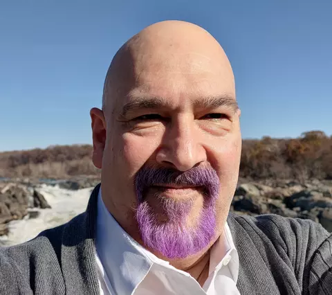 Headshot of a bald man with a purple goatee taking a selfie in front of an outdoor waterfall 