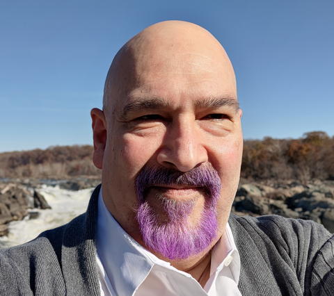 Headshot of a bald man with a purple goatee taking a selfie in front of an outdoor waterfall 