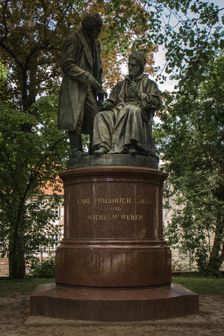 A stone monument on a circular pedestal set in a leafy park shows two men, one seated and one standing. 