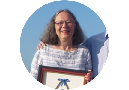 A woman with medium length grey hair wearing a striped shirt, standing outside, holding an award.