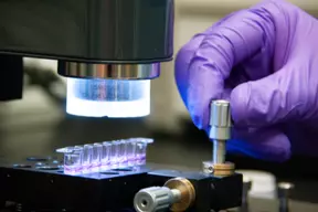 A hand in a purple glove turns a metal dial on a plate holding tiny vials of purple liquid. 