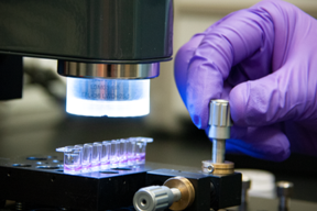 A hand in a purple glove turns a metal dial on a plate holding tiny vials of purple liquid. 