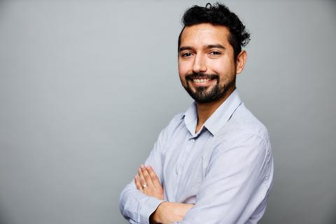 Portrait of Carlos Renteria, a man with short dark hair and a beard, wearing a button up shirt. His arms are folded and he is smiling for the photo.