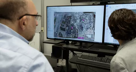 View from behind shows two seated researchers looking at a computer screen that shows an aerial view of a large outdoor site. 
