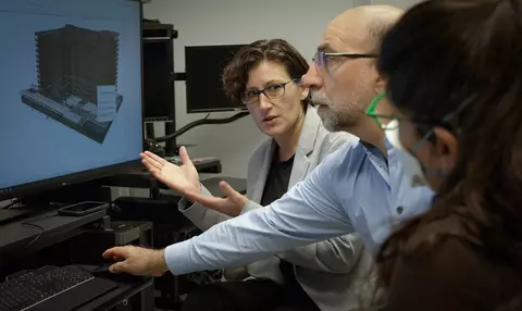 A woman gestures to a building model on a computer screen as she speaks to the two other researchers sitting near her. 